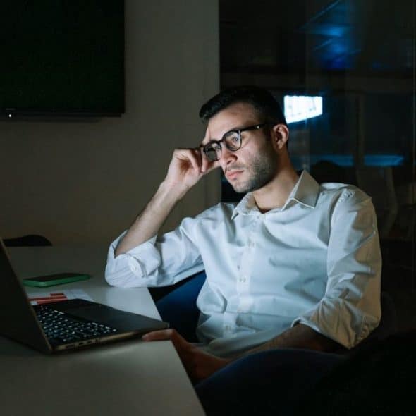 A man in glasses and a white shirt works alone on a laptop in a dark office.