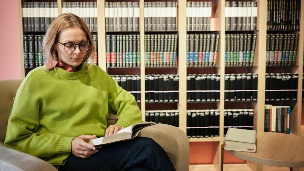 A woman wearing glasses and reading a book while sitting on a couch in a library.