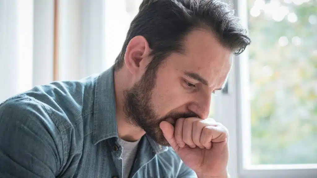 A close-up of a bearded man in a denim shirt looking down, with his fist to his mouth.