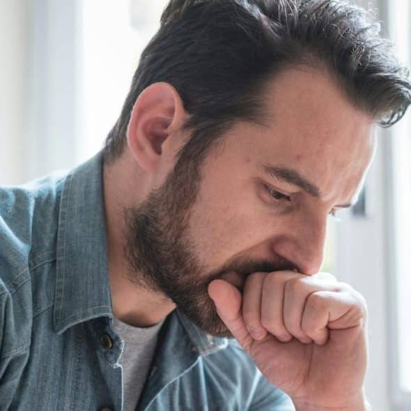 A close-up of a bearded man in a denim shirt looking down, with his fist to his mouth.