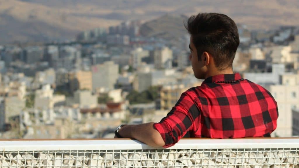 A man wearing a red plaid shirt sitting on a bench overlooking an urban area.
