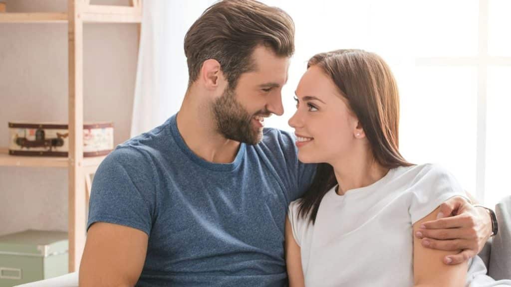 Man with arm around woman, both smiling and looking at each other indoors.