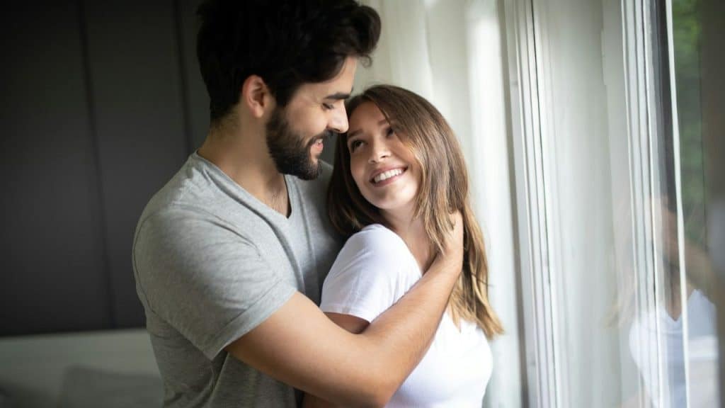 Man and woman stand smiling by a window, embracing and looking at each other.