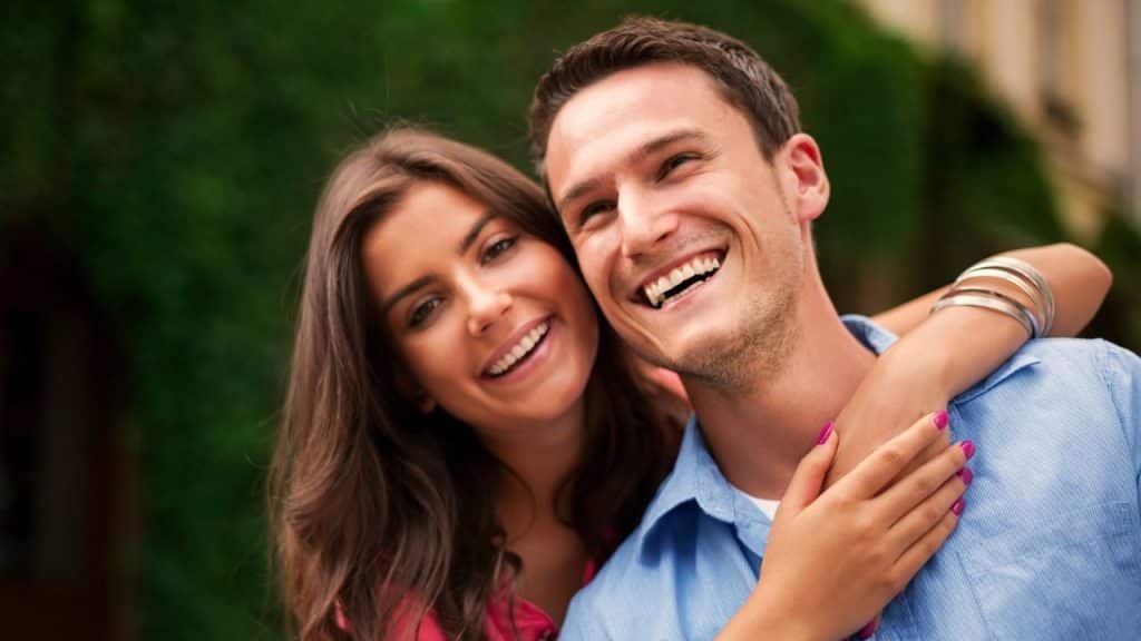 Smiling man in a blue shirt with a woman hugging him from behind.