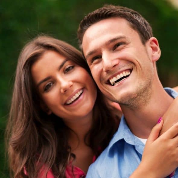 Smiling man in a blue shirt with a woman hugging him from behind.