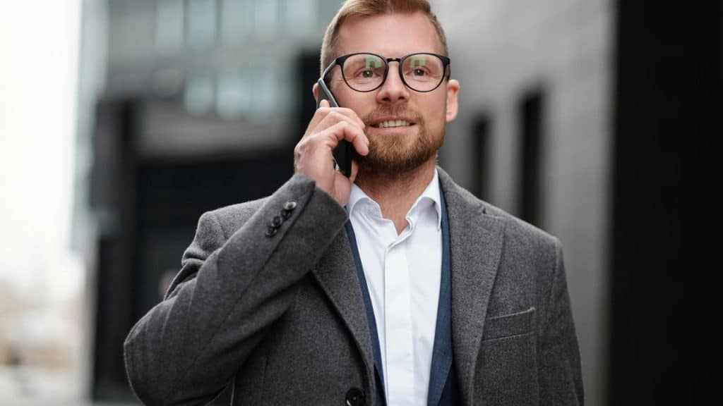 A man in glasses and a grey coat talking on a phone outdoors.