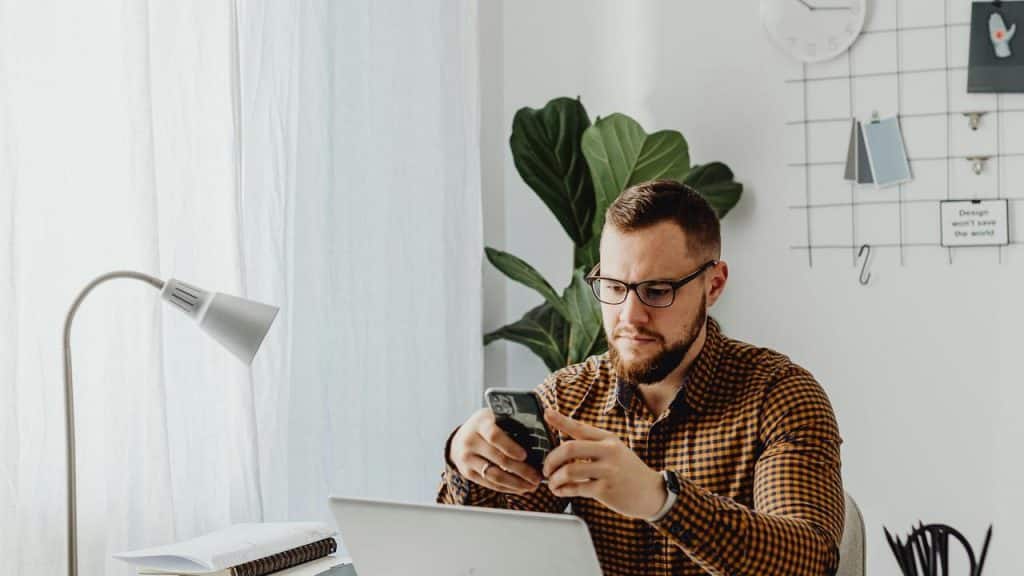 A man using a smartphone at a desk with a laptop.