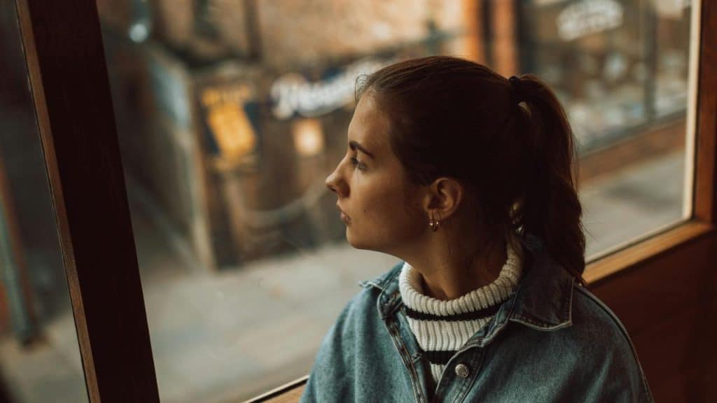 A young woman in a denim jacket looking out a window with a thoughtful expression.