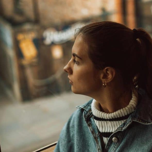 A young woman in a denim jacket looking out a window with a thoughtful expression.