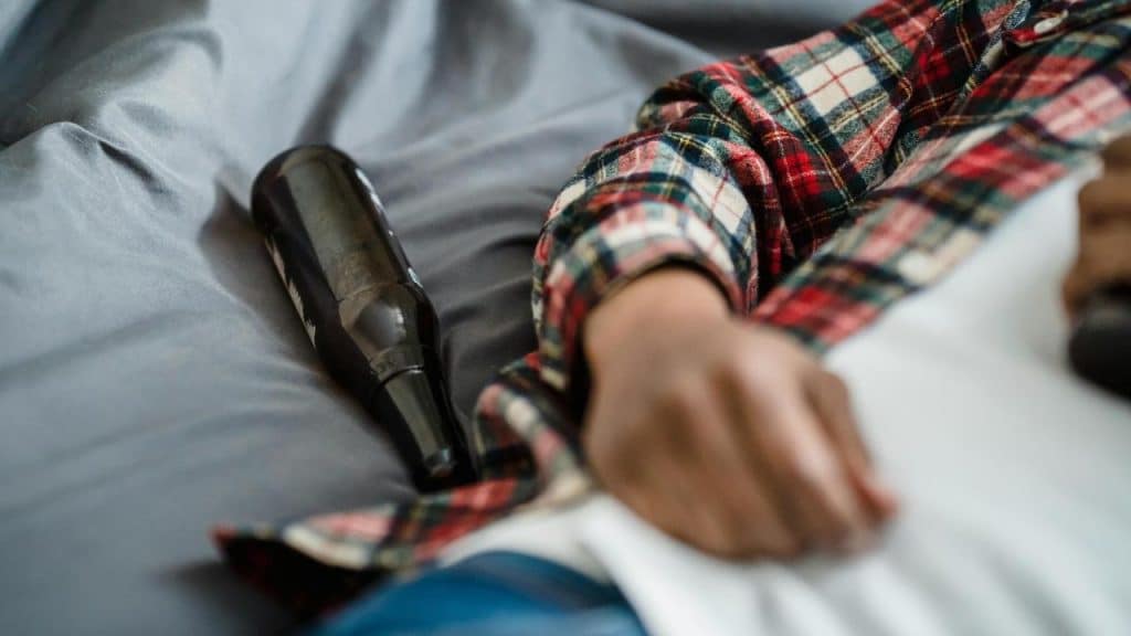 A person’s hand resting on a bed beside an empty bottle.
