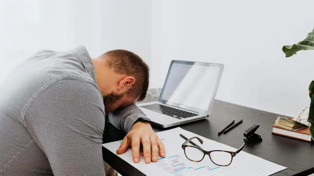 A man resting his head on a desk beside a laptop and papers.
