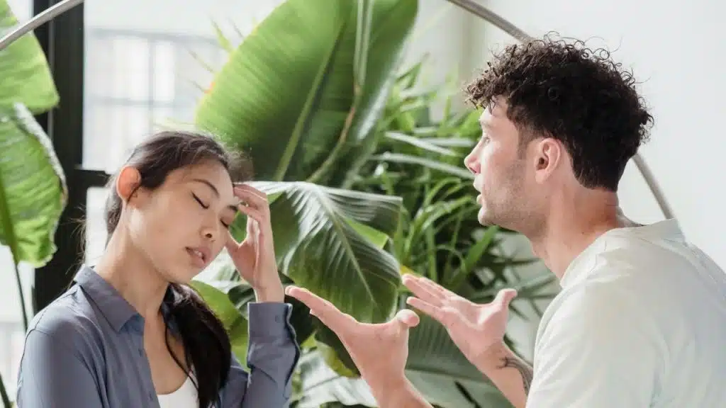 A man is passionately arguing, gesturing with his hands, while a woman beside him looks distressed, holding her head.