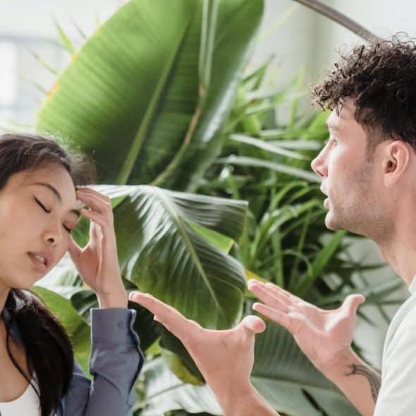 A man is passionately arguing, gesturing with his hands, while a woman beside him looks distressed, holding her head.
