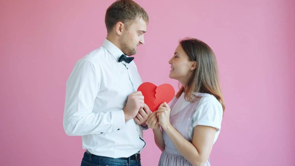 A couple holding a broken heart on a pink background.