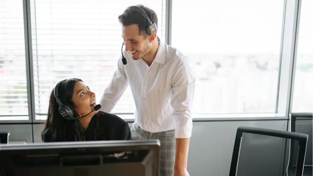 Two call center employees wearing headsets are smiling and looking at each other over a desk with a computer monitor.