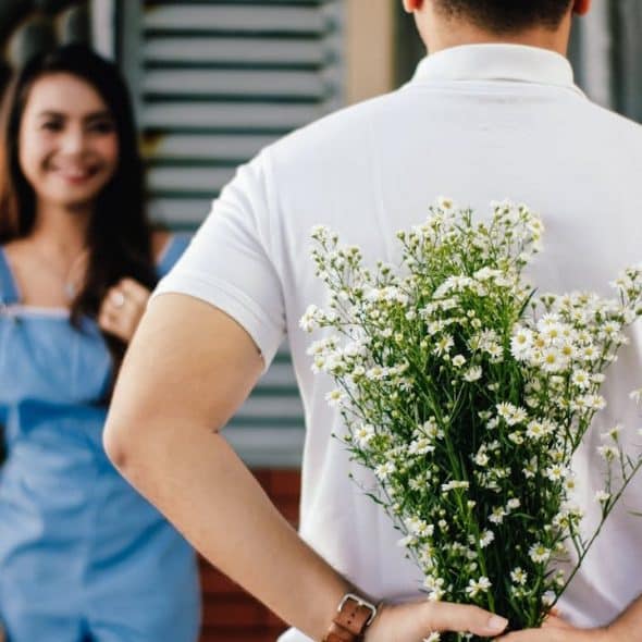 A man stands with his back to the camera, holding a bouquet of white flowers behind him, while a smiling woman in a blue outfit stands blurred in the background.