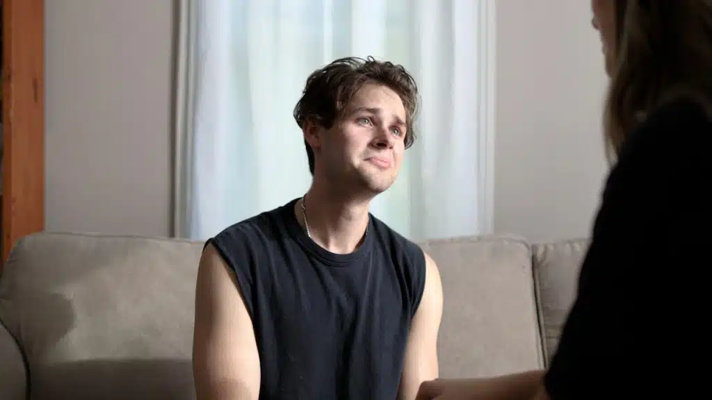 A distressed young man is sitting on a sofa, looking up at a woman in the foreground with a sad expression.