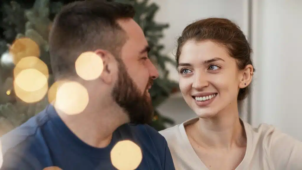 A happy couple is looking and smiling at each other, with festive lights blurring in the foreground.