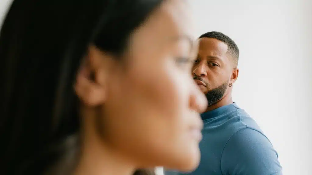 A close-up shot of a Black man in a blue shirt staring directly at the camera over the shoulder of a blurred woman in the foreground.