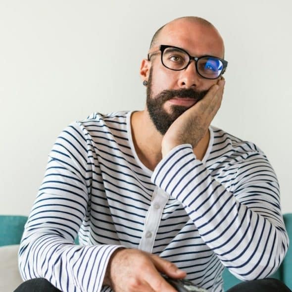 A bald, bearded man wearing glasses sitting on a couch and looking frustrated.