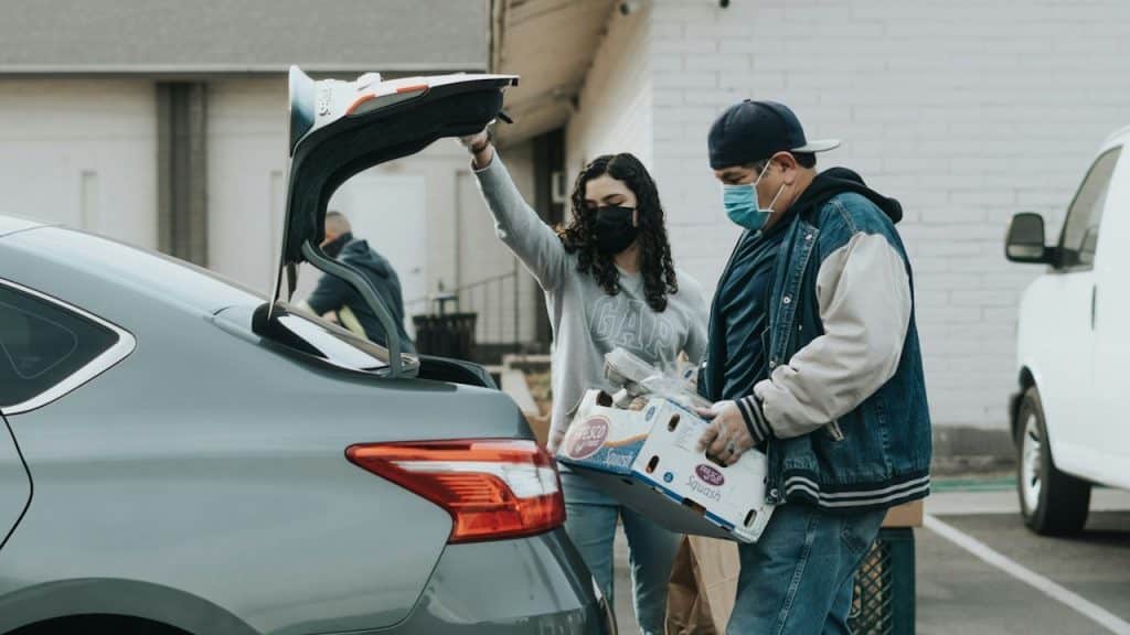 Two people wearing face masks are loading boxes of food into the open trunk of a gray car