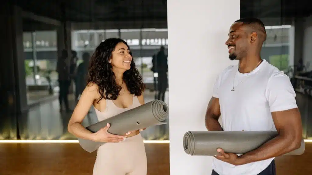 A man and a woman in workout clothes are standing, holding yoga mats, and smiling at each other in a fitness studio.