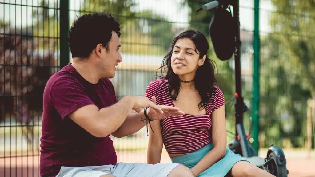 A man and woman are sitting outdoors near a fence and a scooter, talking and looking engaged in conversation.