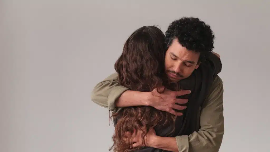 Man with a mustache hugging a woman from behind with a serious expression against a plain background.