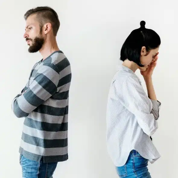 Couple standing with their backs to each other and the women is looking upset while holding her hand up to her face.