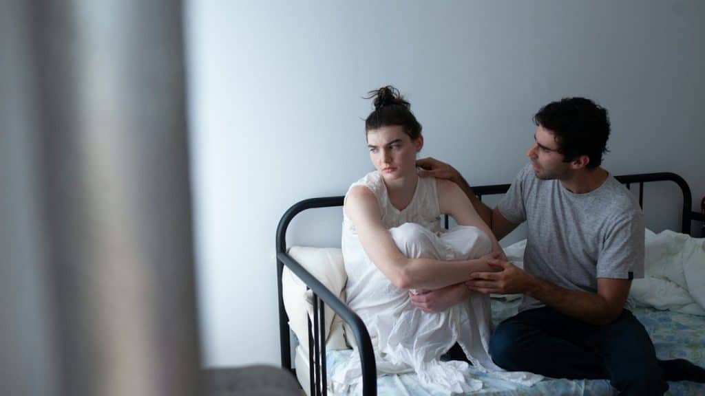 A man comforting a distressed woman sitting on a bed.