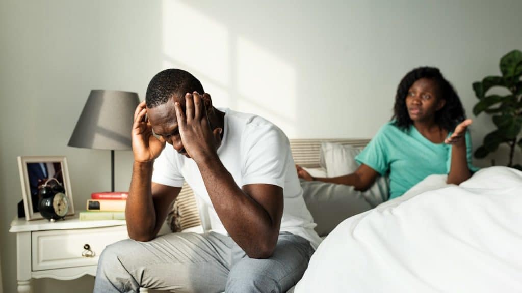 A distressed Black man sits on the edge of a bed, holding his head, while a Black woman argues with him in the background.