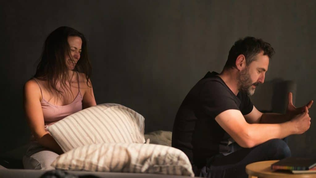 A woman is crying and sitting on a bed with a pillow while a man sits beside her looking distressed and away from her.