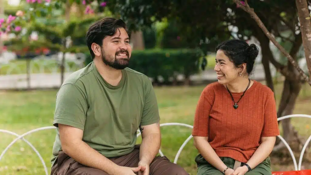 A smiling couple is sitting outdoors on a red bench, facing and looking happily at each other under a flowering tree.