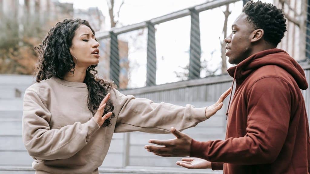 A woman with curly hair pushes away a man in a hoodie outside.