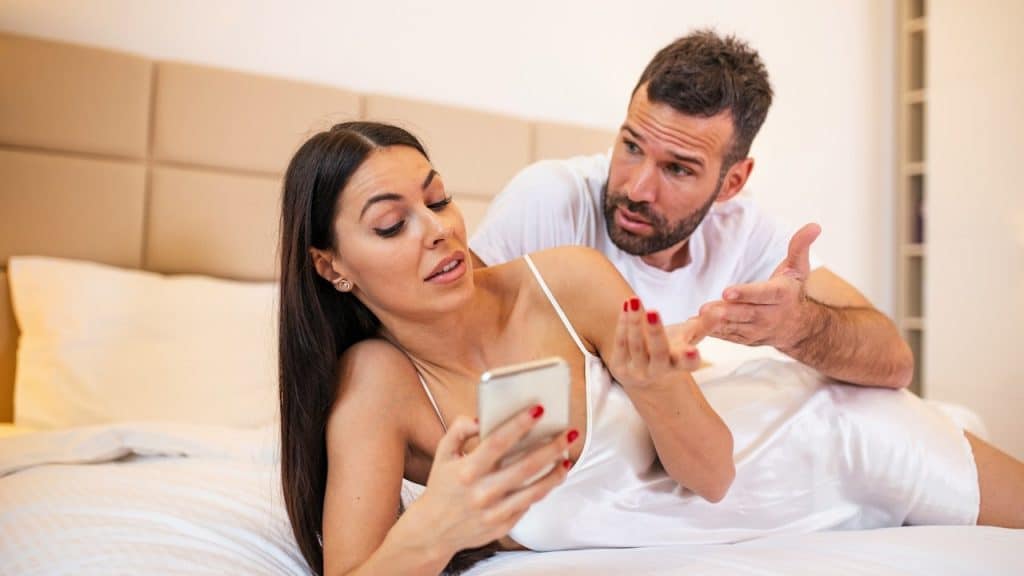 A man looking frustrated and gesturing with his hands towards a woman who is ignoring him while looking at her phone in bed.