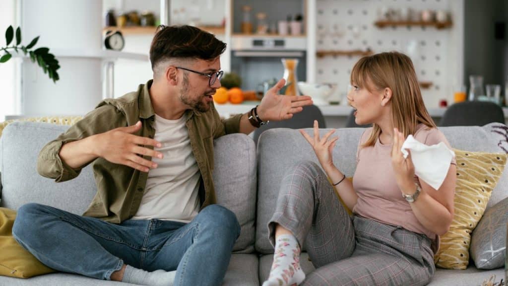 A couple arguing on a gray couch, both gesturing with their hands while looking at each other.