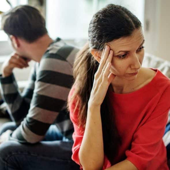 A distressed woman in a red top sits in the foreground, facing away from a man.