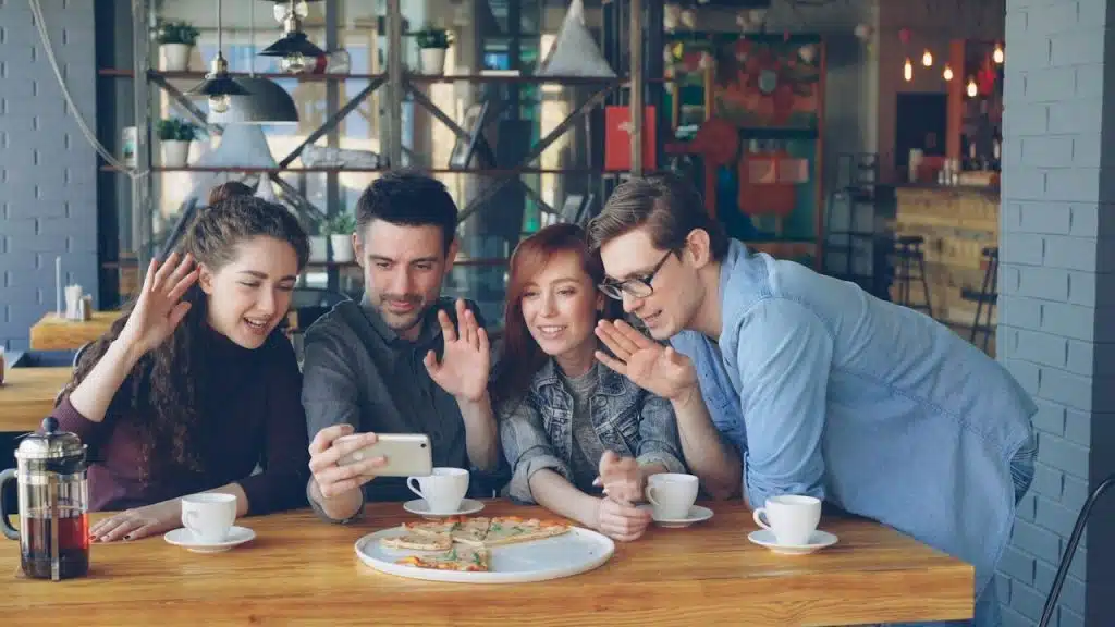 Four friends are sitting around a table in a cafe with pizza and coffee, waving at a mobile phone, likely on a video call.