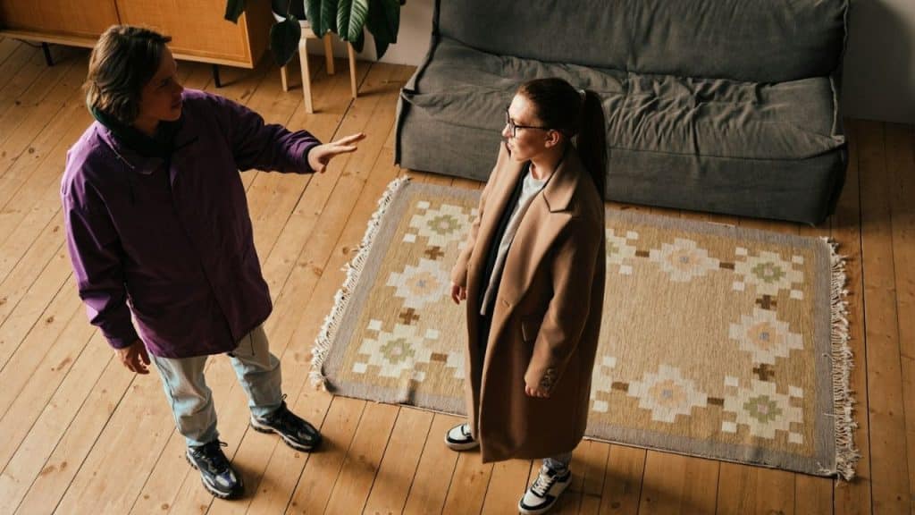 Two people in coats arguing in a living room, one has hand raised.