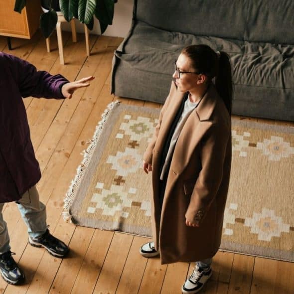 Two people in coats arguing in a living room, one has hand raised.