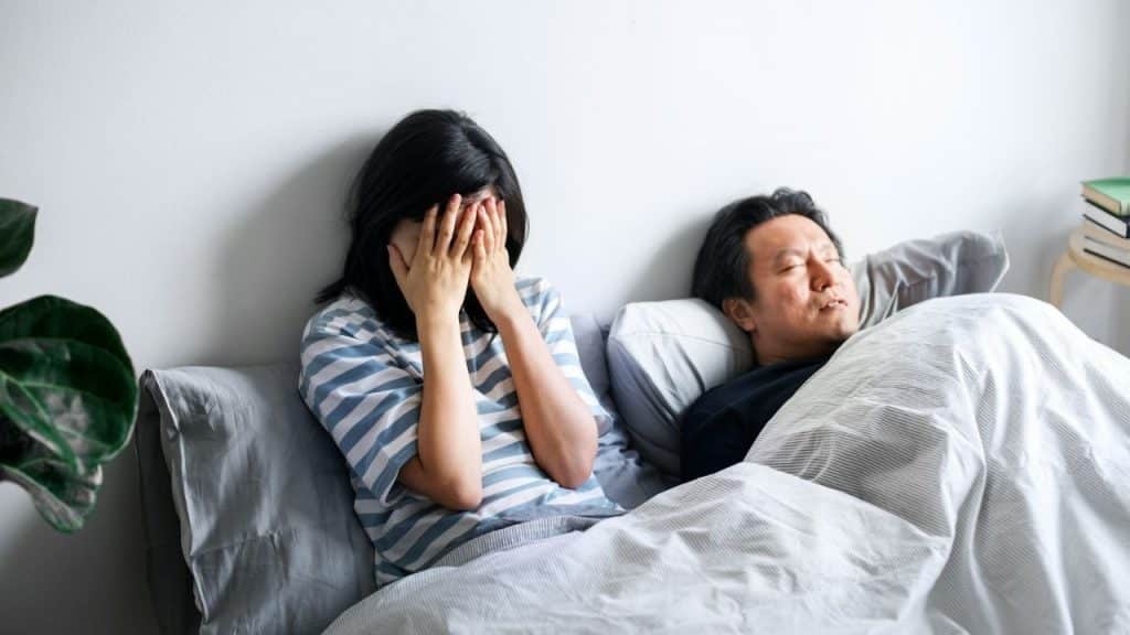 A woman is sitting up in bed, covering her face; a man sleeps next to her.