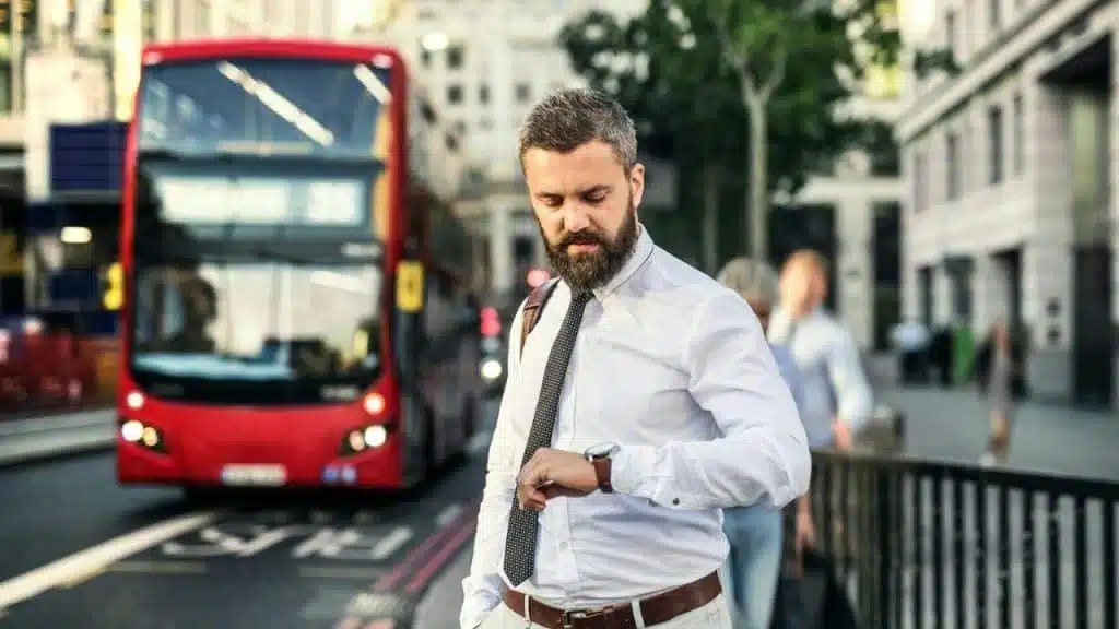 Businessman checking his watch on a street with a red double-decker bus behind him.