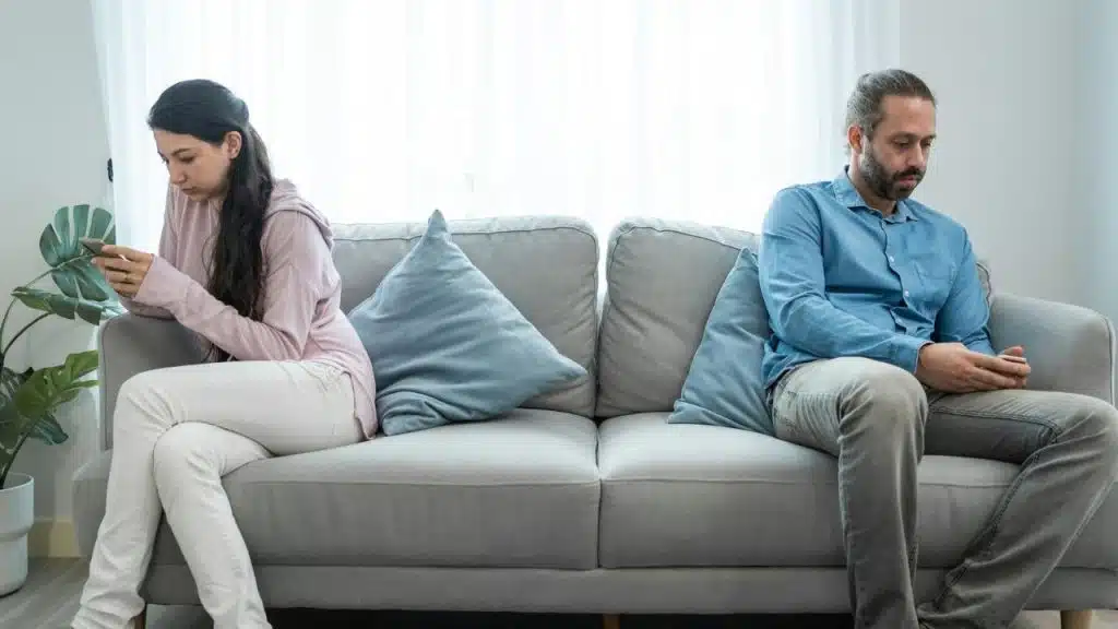 Man and woman sitting far apart on a gray couch, ignoring each other.