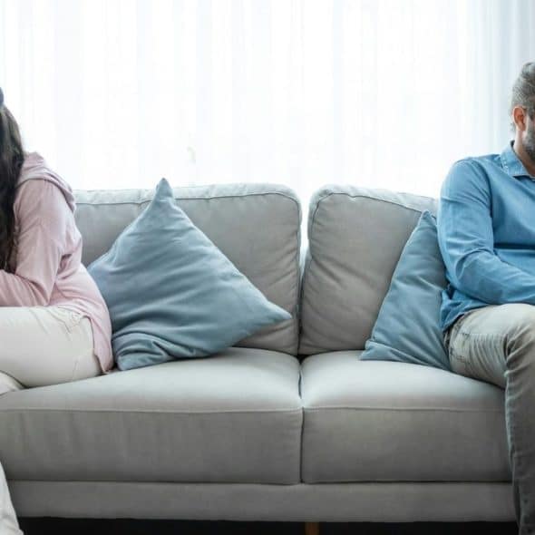Man and woman sitting far apart on a gray couch, ignoring each other.