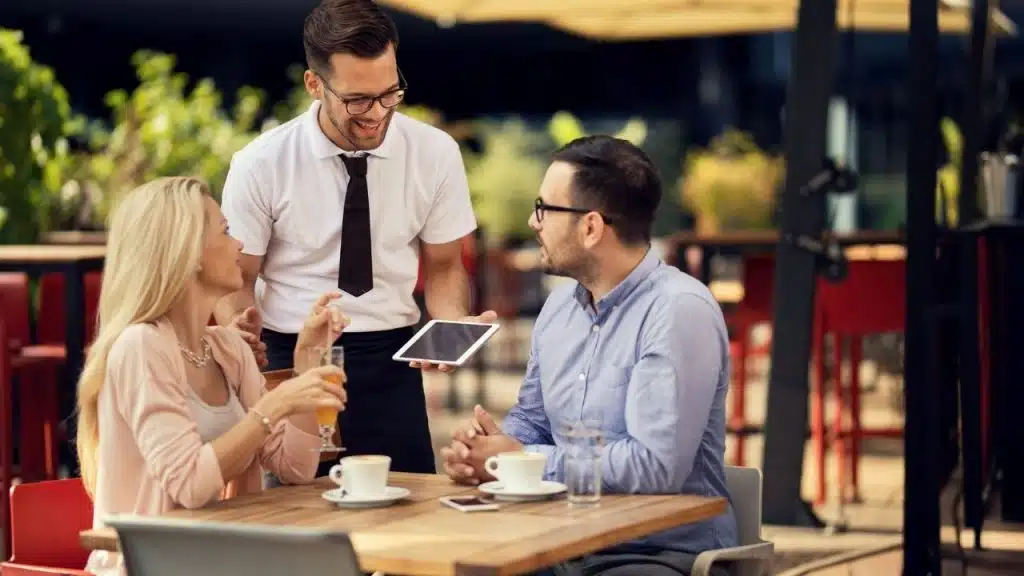 Smiling waiter showing a digital tablet to a couple seated at an outdoor cafe table.
