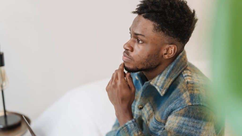 A thoughtful young Black man in a blue and yellow plaid jacket is sitting with his hand on his chin, looking away.