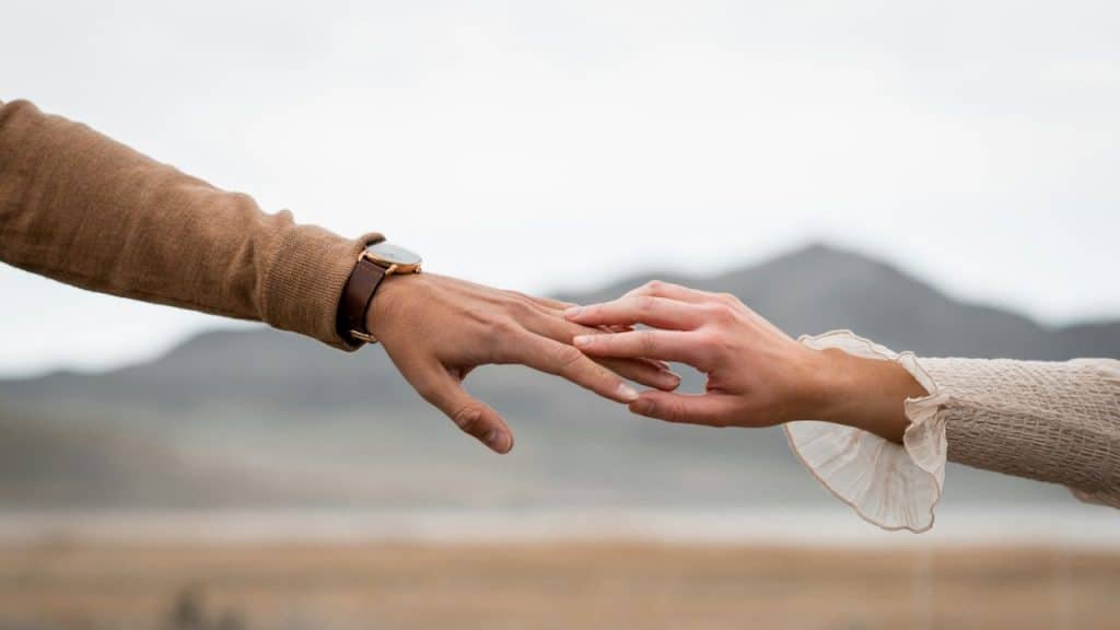 A close-up of a man's and a woman's hands gently touching fingers, with a blurred natural landscape and mountains in the background.