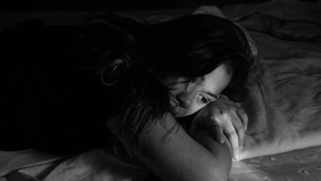 A black and white image of a woman lying on a bed with her head resting on her arms, looking sad or troubled.