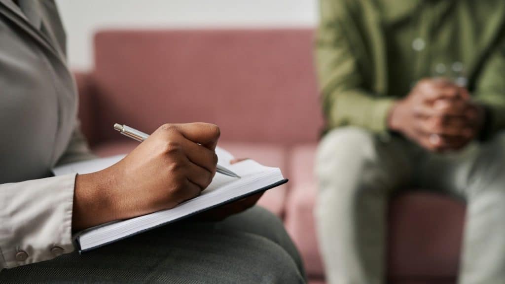 A close-up shot of a therapist or counselor's hands writing notes in a notebook, with a male patient sitting across from them in the blurred background.