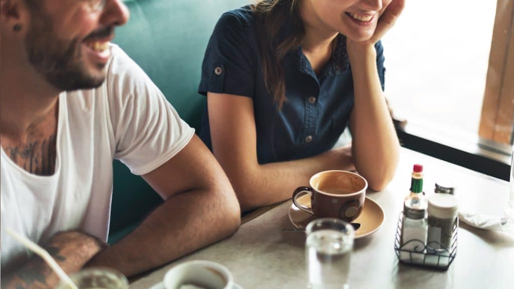 A happy couple is sitting at a cafe table, with the woman smiling and resting her chin on her hand, and the man laughing.